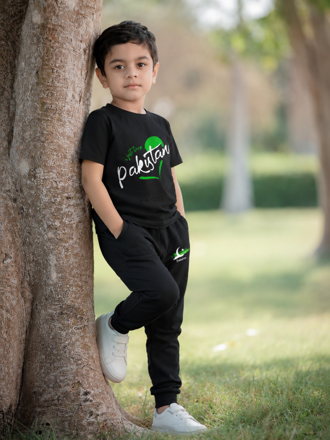 A young boy wearing a black t-shirt and matching sweatpants with green and white 'Pakistan' graphics, leaning against a tree in a park.