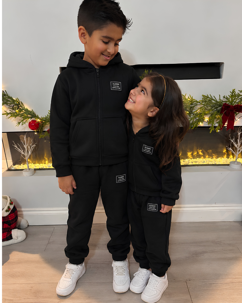 Two children wearing matching black zip-up hoodies and jogger sweatpants stands in a holiday-themed room in front of a fireplace. The boy is standing taller and looking down at the girl, who is smiling up at him.
